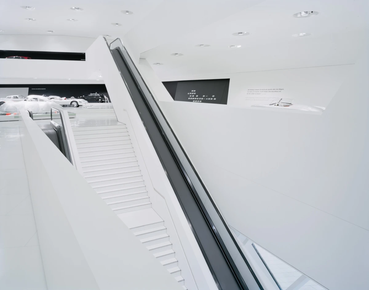 Ascending white escalator beside a staircase in a minimalist car showroom; cars on display in the background.