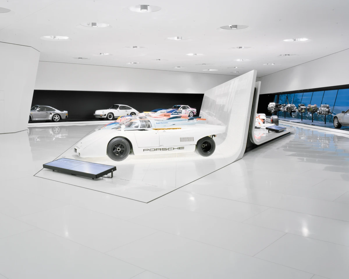 White Porsche race car on a display platform in a bright, minimalist showroom; other cars visible in the background.