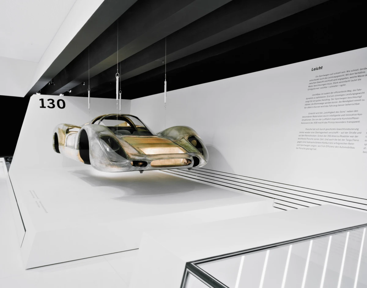 Suspended vintage prototype race car with metal and wood panels, displayed in a white museum alcove labeled 130.