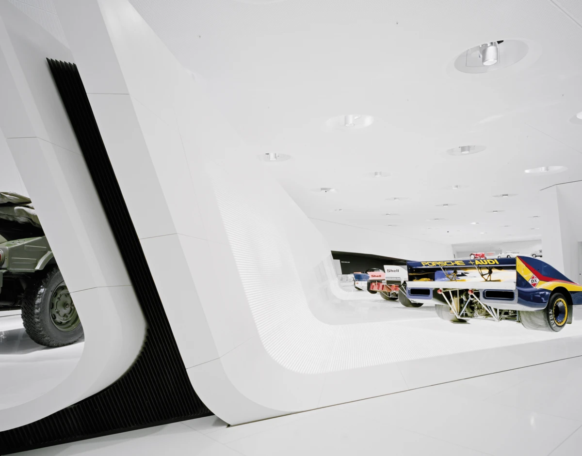 White futuristic showroom with curved walls displaying a blue-yellow Porsche-Audi race car; another race car visible in the background.