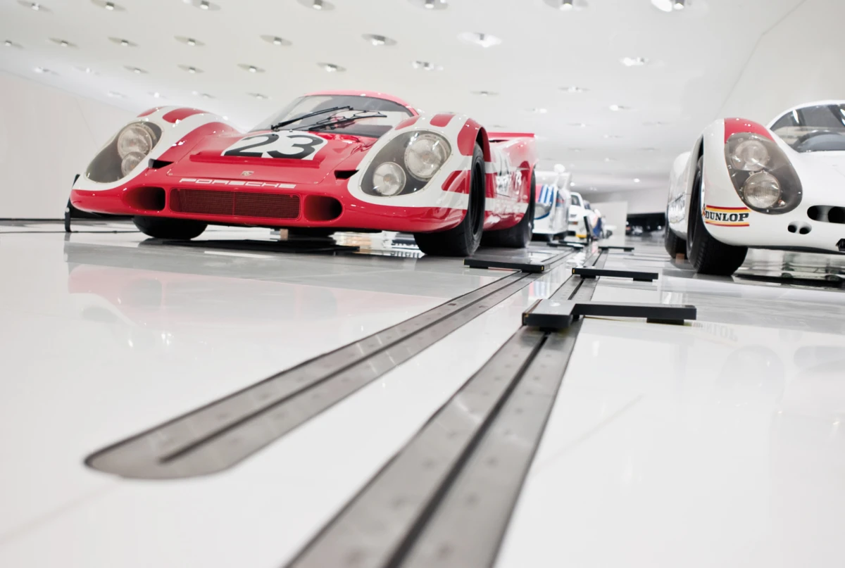 Red Porsche race car with number 23 parked on a glossy showroom floor beside other vintage race cars.