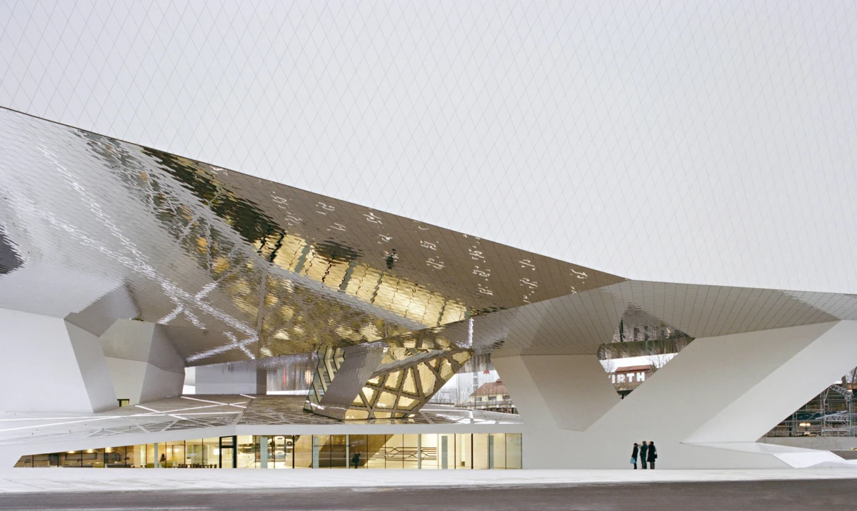 Geometric modern building with a gold reflective roof and white angular supports; two people stand in a plaza.