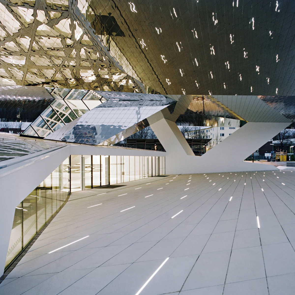 Futuristic glass pavilion with white angular supports and mirrored roof; outdoor plaza with illuminated walkway.