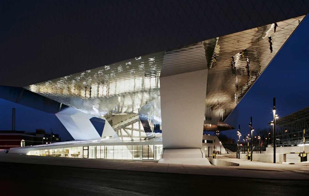 Futuristic glass-and-steel building with a slanted, reflective canopy illuminated at night beside a lit street.