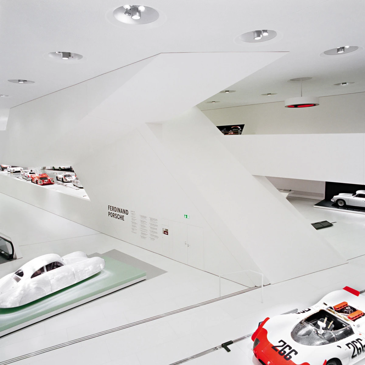 White modern museum gallery displaying Porsche race-car models on raised platforms; wall reads Ferdinand Porsche.