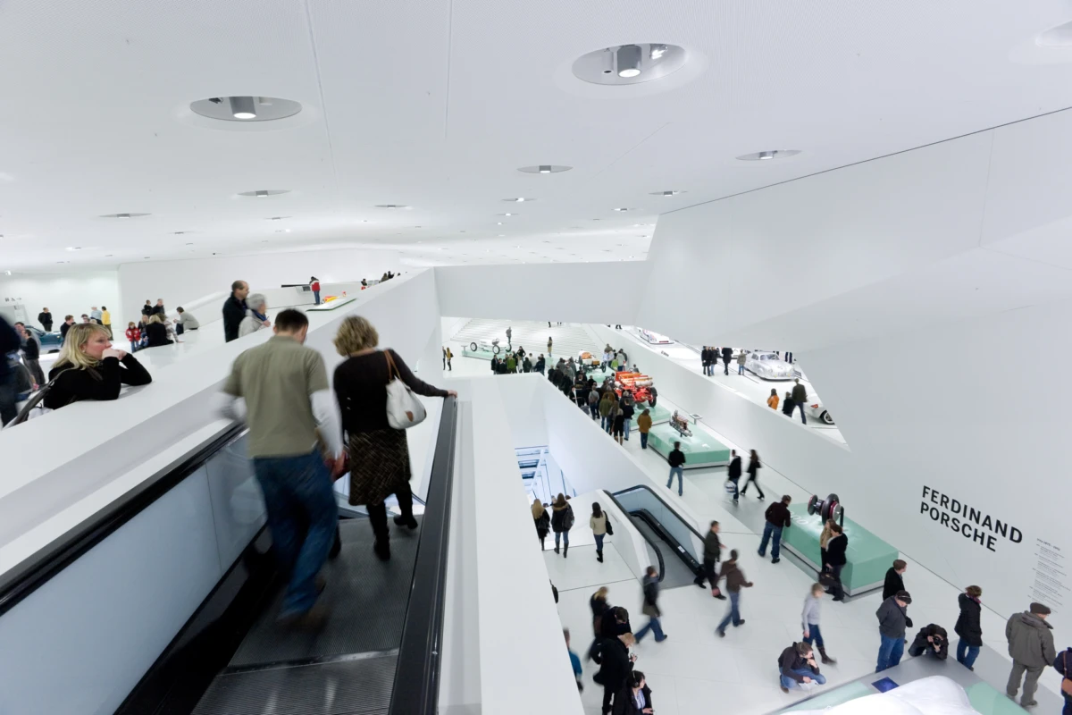 Visitors ride an escalator and walk through a bright white Porsche museum, with FERDINAND PORSCHE signage on the wall.