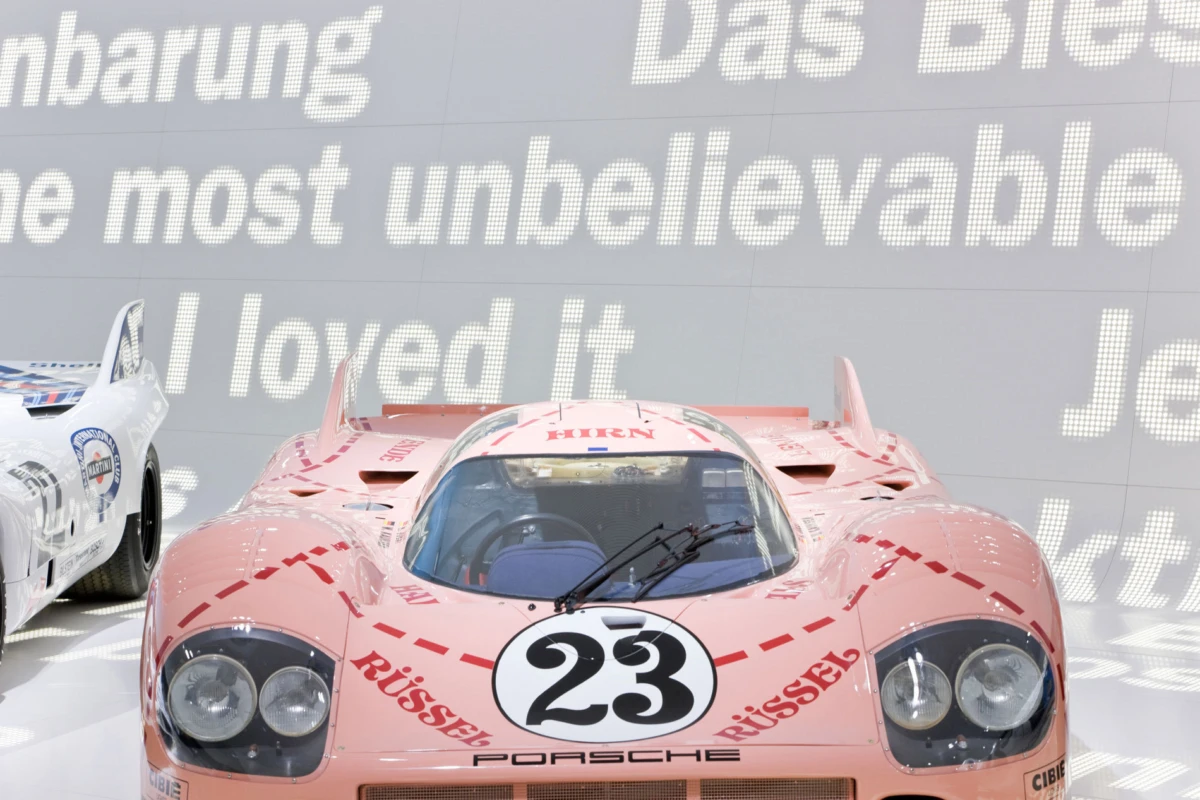 Pink Porsche race car, number 23, on display in a showroom; white race car visible to the left.