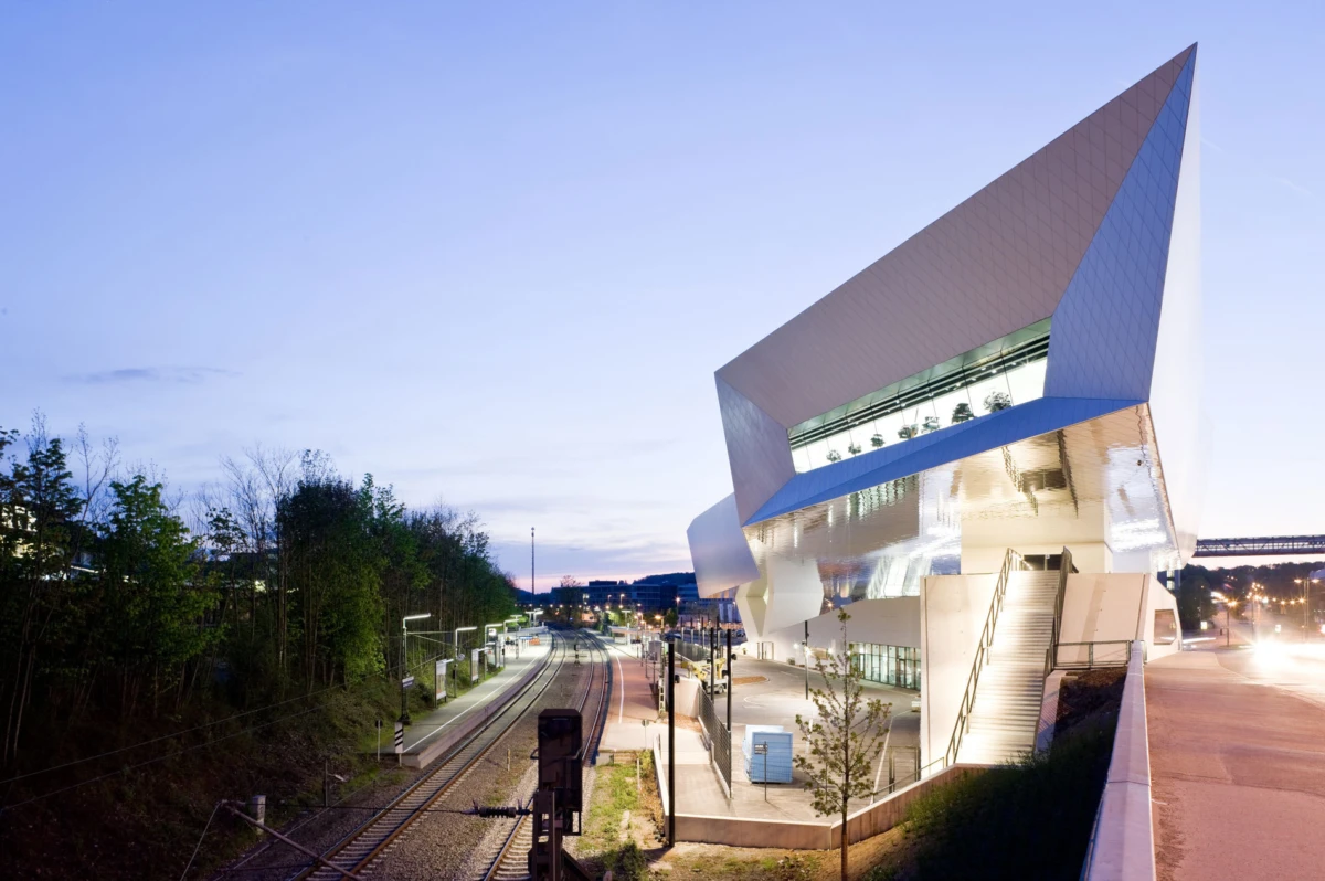 Contemporary angular white building with glass facade beside railway tracks and a road at dusk; exterior stairs lead to the entrance.