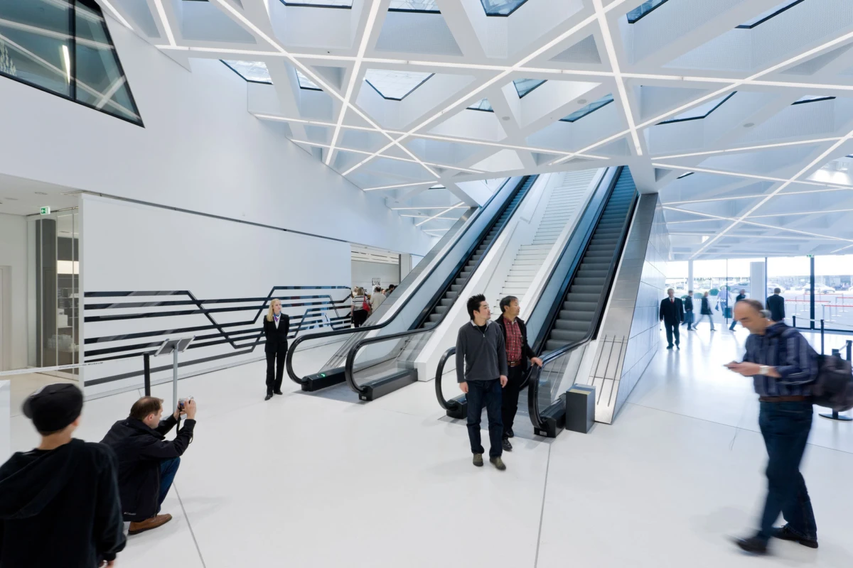 Two people stand by a pair of escalators in a bright white futuristic atrium with a geometric ceiling; others walk by.