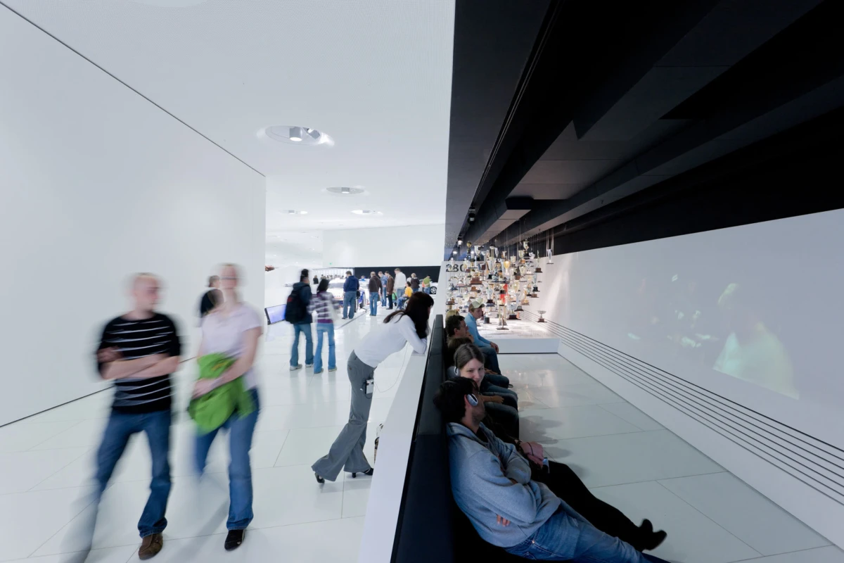Bright white contemporary art gallery corridor with visitors walking; some sit along a bench beside a wall displaying sculptures.