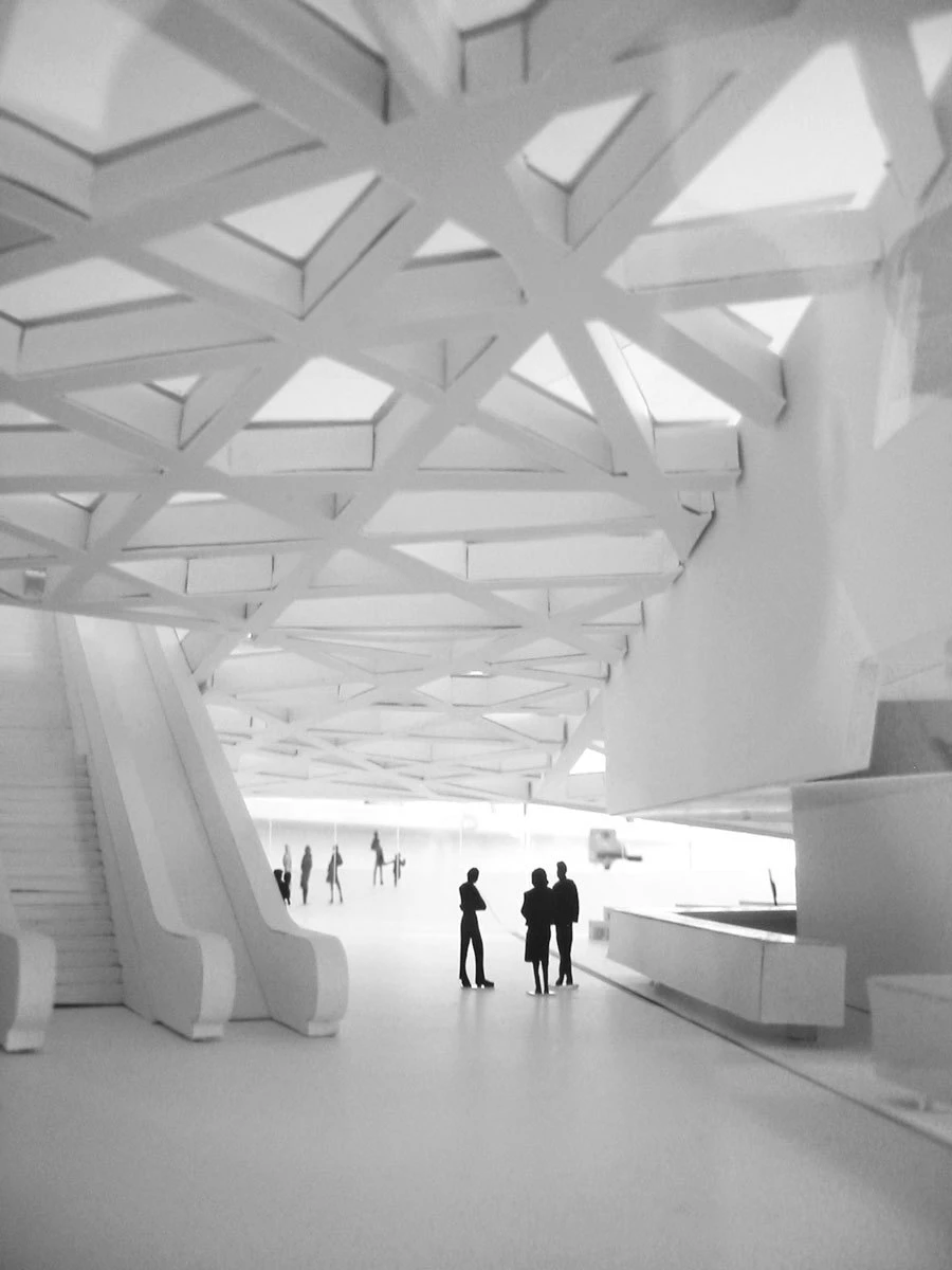 Interior of a white modern building with a crisscross lattice ceiling; silhouetted groups of people standing and chatting.