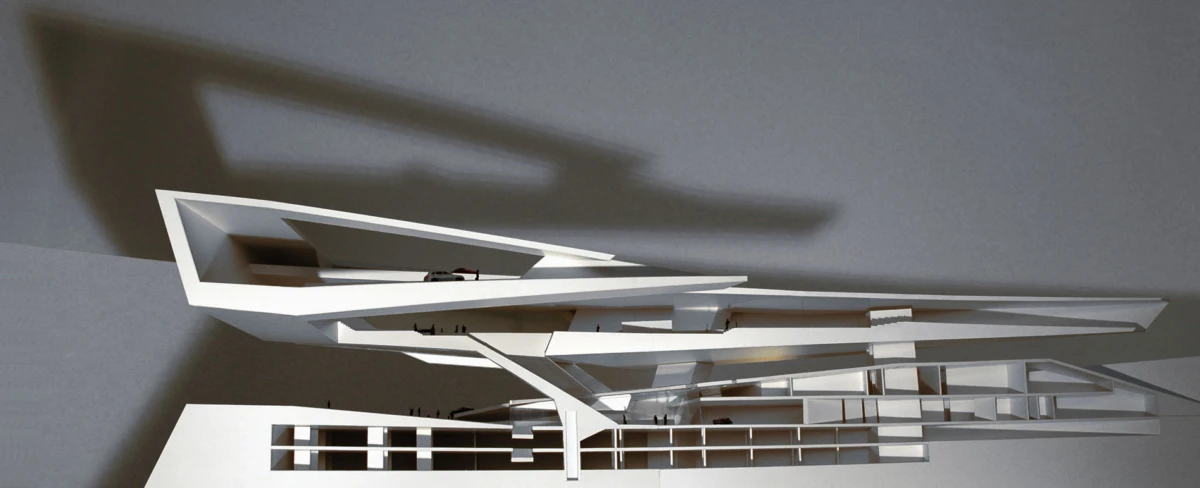 White angular architectural model with interlocking beams, displayed indoors against a gray wall casting shadows.