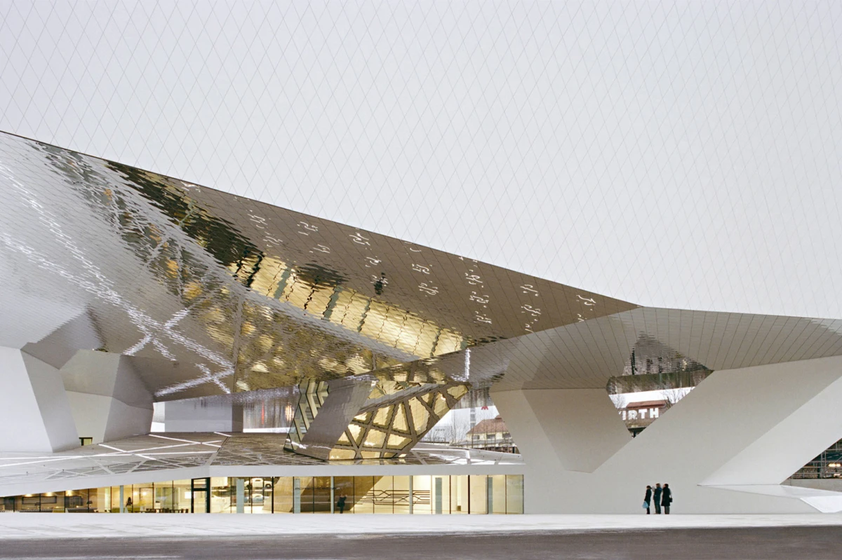 Futuristic white, angular building with reflective metallic panels; two people stand at its base.