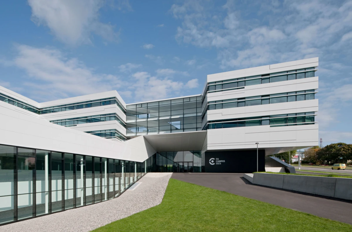 Modern white campus building with glass facade and angled design; curved entrance, green lawn, FH Campus Wien sign.