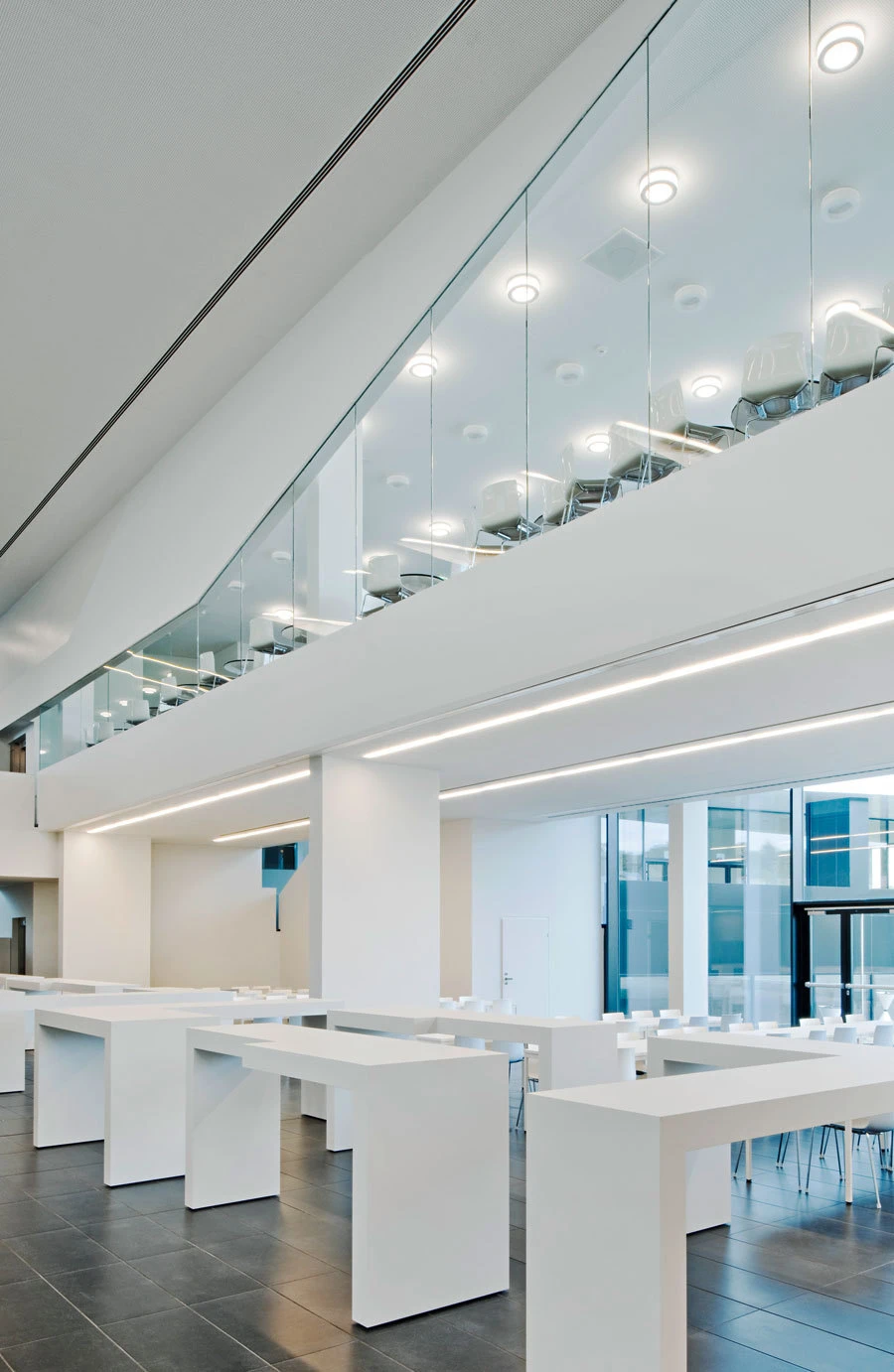 Minimalist white interior with a glass-railed mezzanine and rows of white high-top tables in an open workspace.
