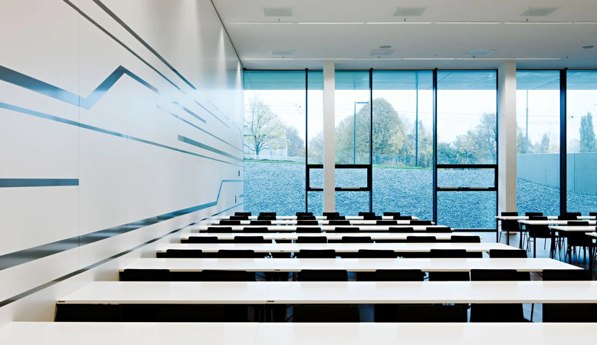 Empty modern lecture hall with rows of white desks and black chairs facing large glass windows.