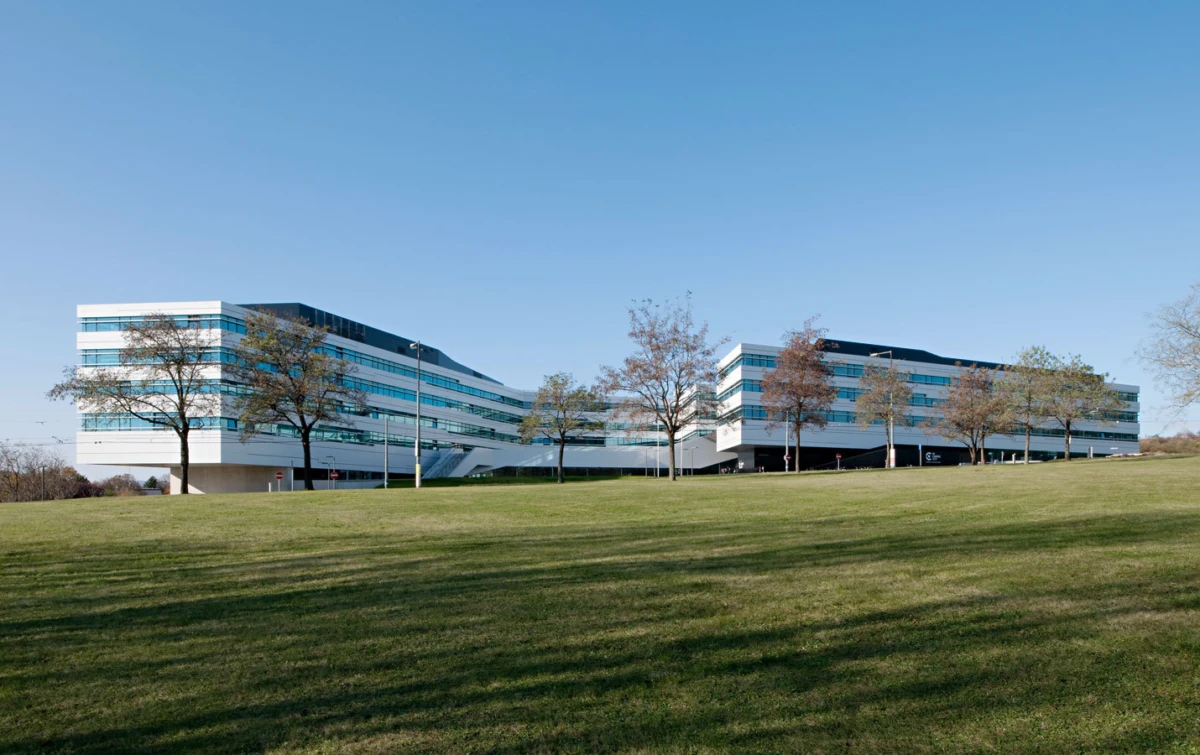 Two low, elongated glass office buildings connected by a central hub, set on a grassy lawn with trees under a clear blue sky.