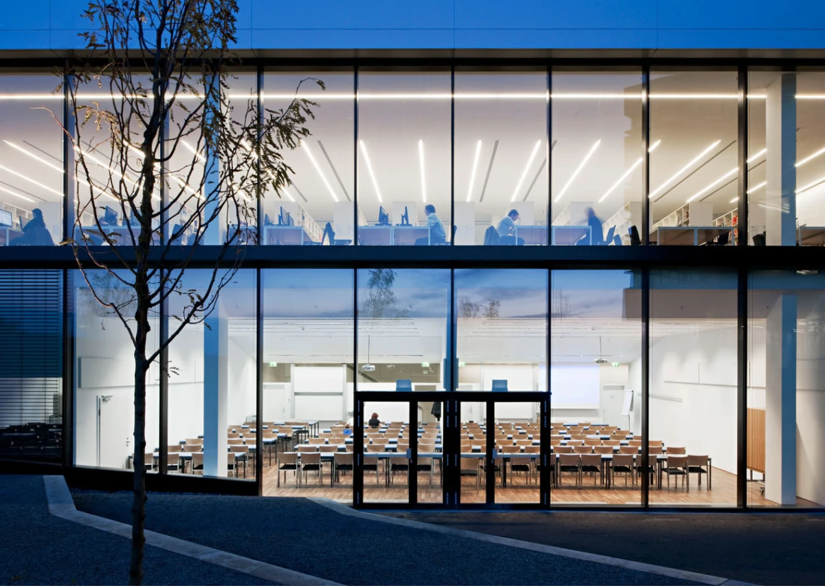Glass-walled building; ground-floor conference room with rows of chairs, upper-floor study area with people at desks under linear lights; bare tree in foreground.