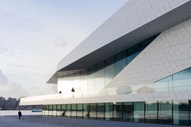 Angular white geometric waterfront building with glass facade; a person stands on a balcony while another walks along the promenade.