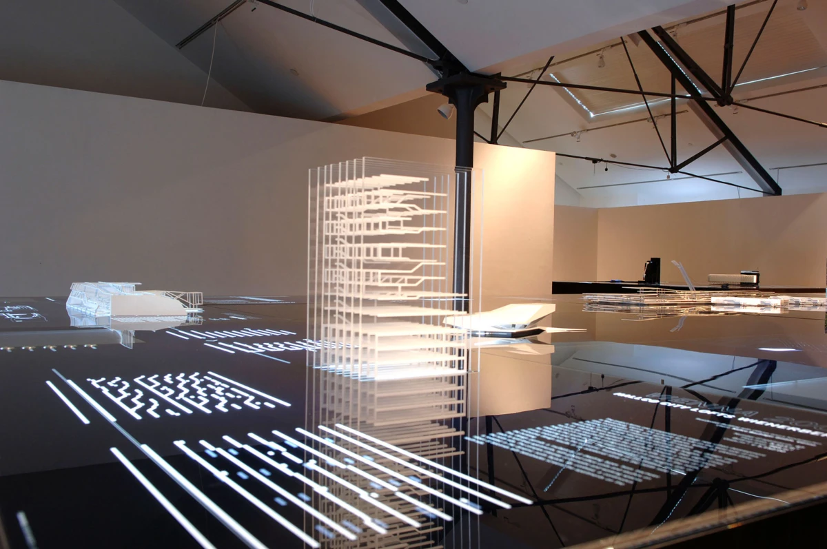 Architectural models and translucent towers on a reflective tabletop in a modern gallery with exposed beams.