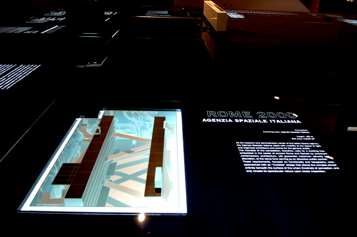 3D architectural model in a glass display case illuminated in a dark exhibition space; text panel reads ROME 2000 by Agenzia Spaziale Italiana.