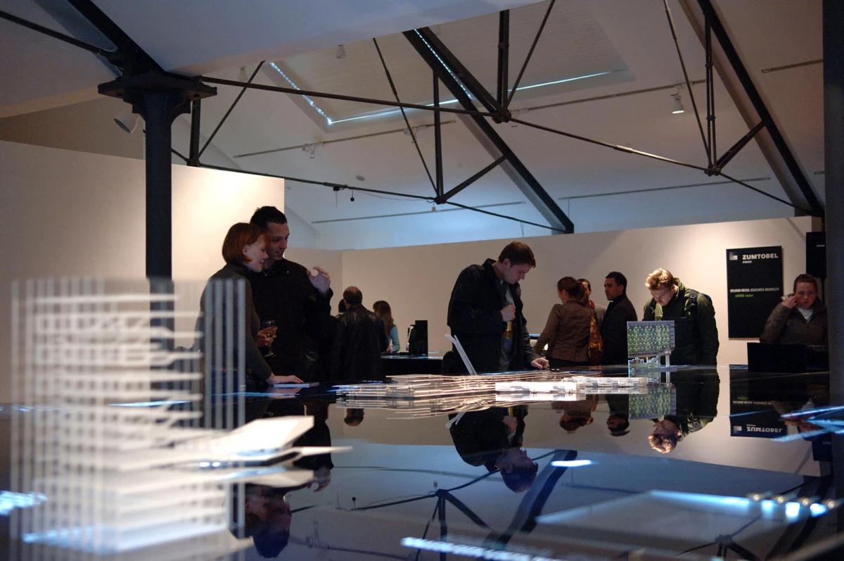 Group of visitors in a modern gallery examining architectural models on a glossy, reflective table.