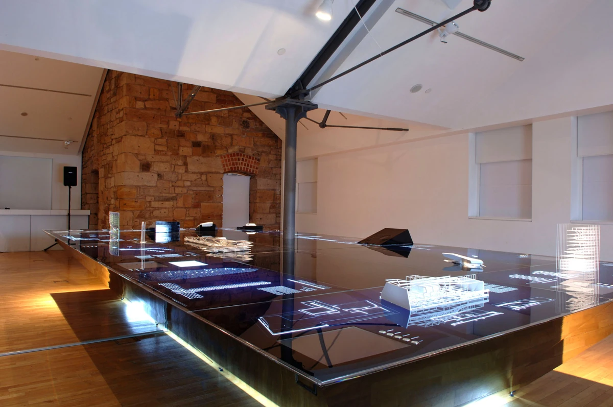 Glass-topped interactive table displaying holographic architectural diagrams and models in a gallery with a stone wall.