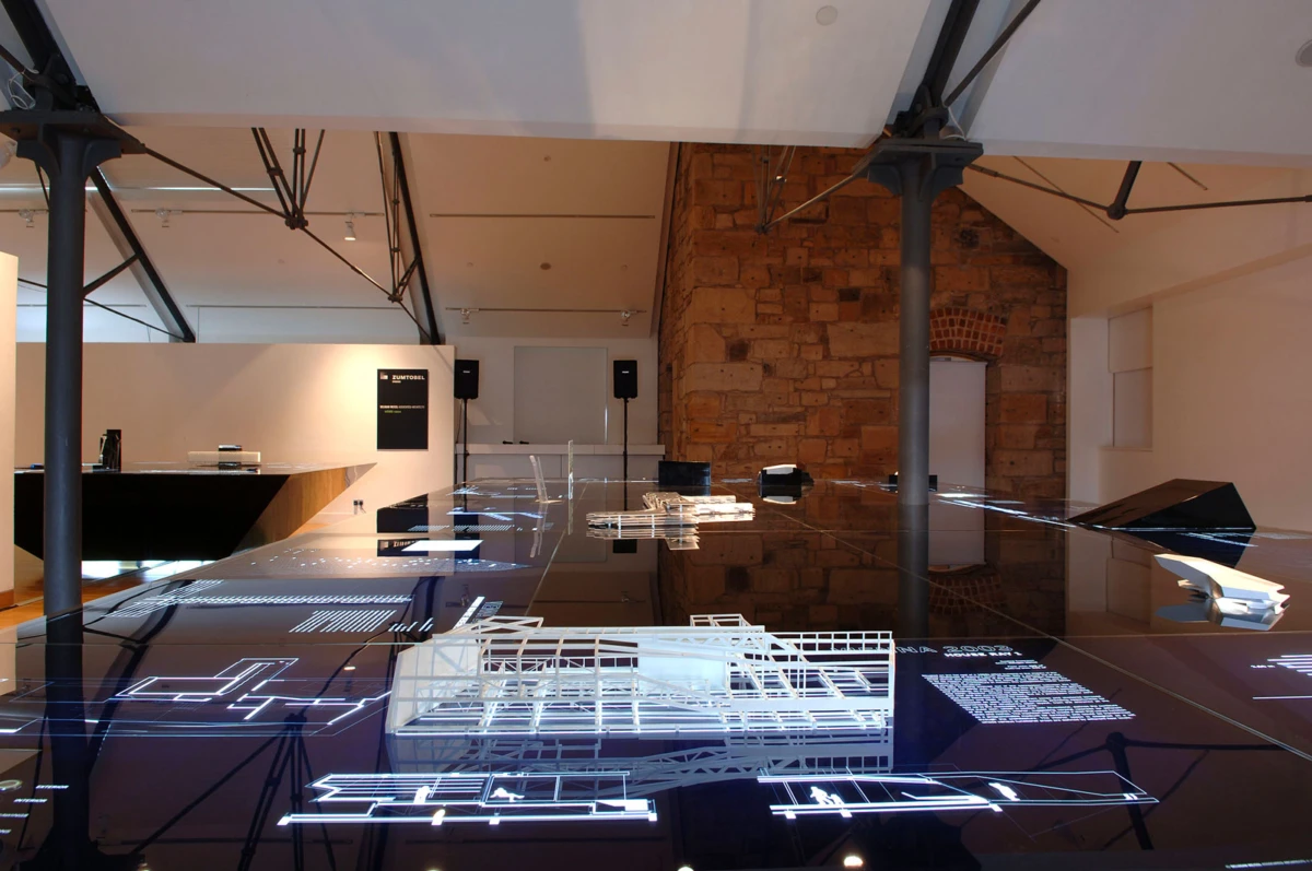 Architectural installation: white line-drawn blueprints displayed on a glossy glass floor in a modern gallery with a brick wall.