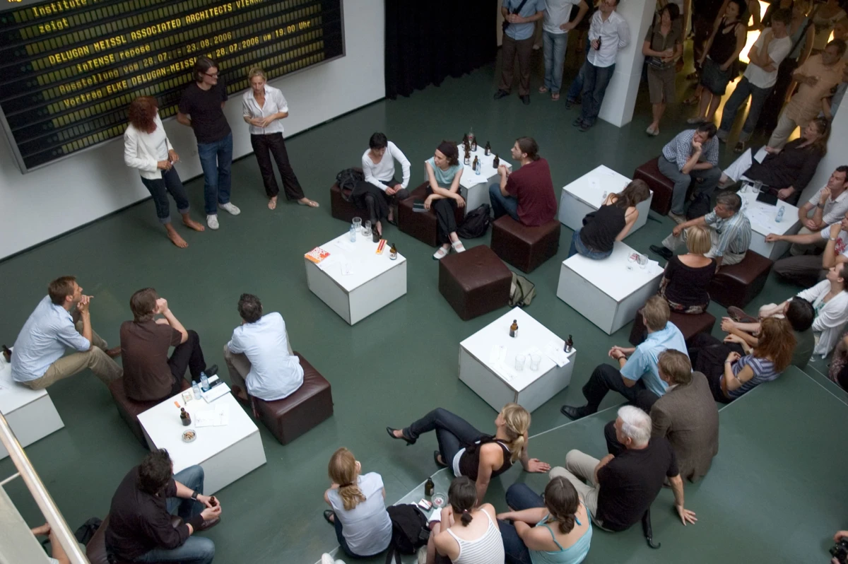 Audience seated on white cube tables and brown ottomans in a contemporary gallery, watching a presentation near a large digital board.