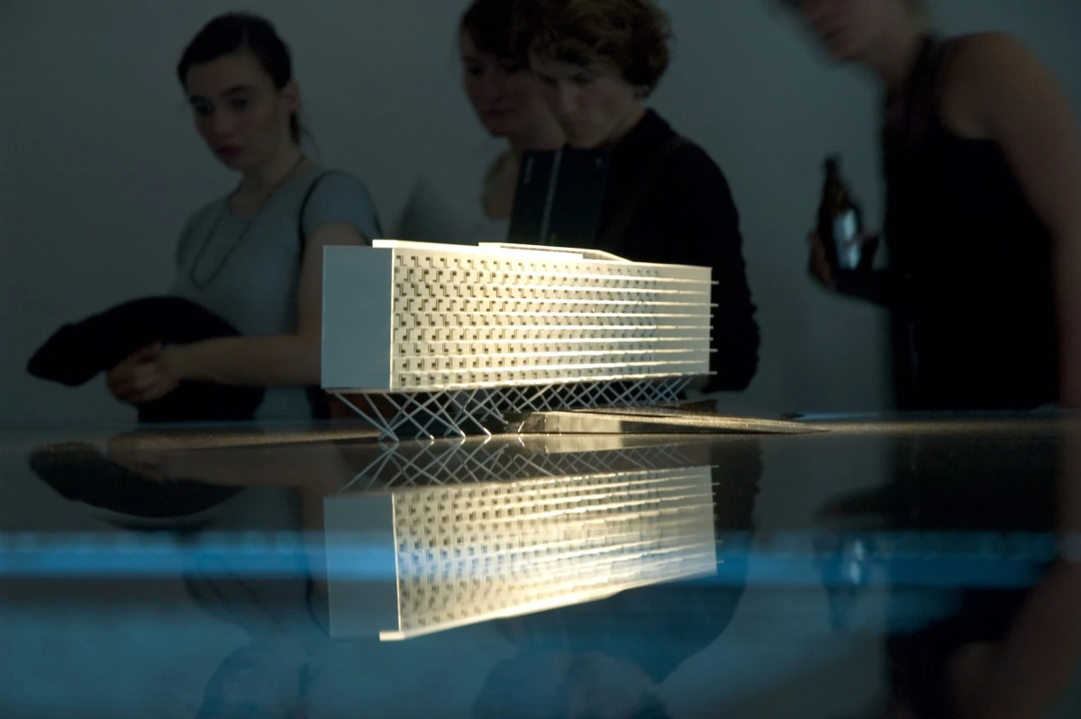 White multi-story architectural model displayed on a reflective table, with blurred visitors in the background.