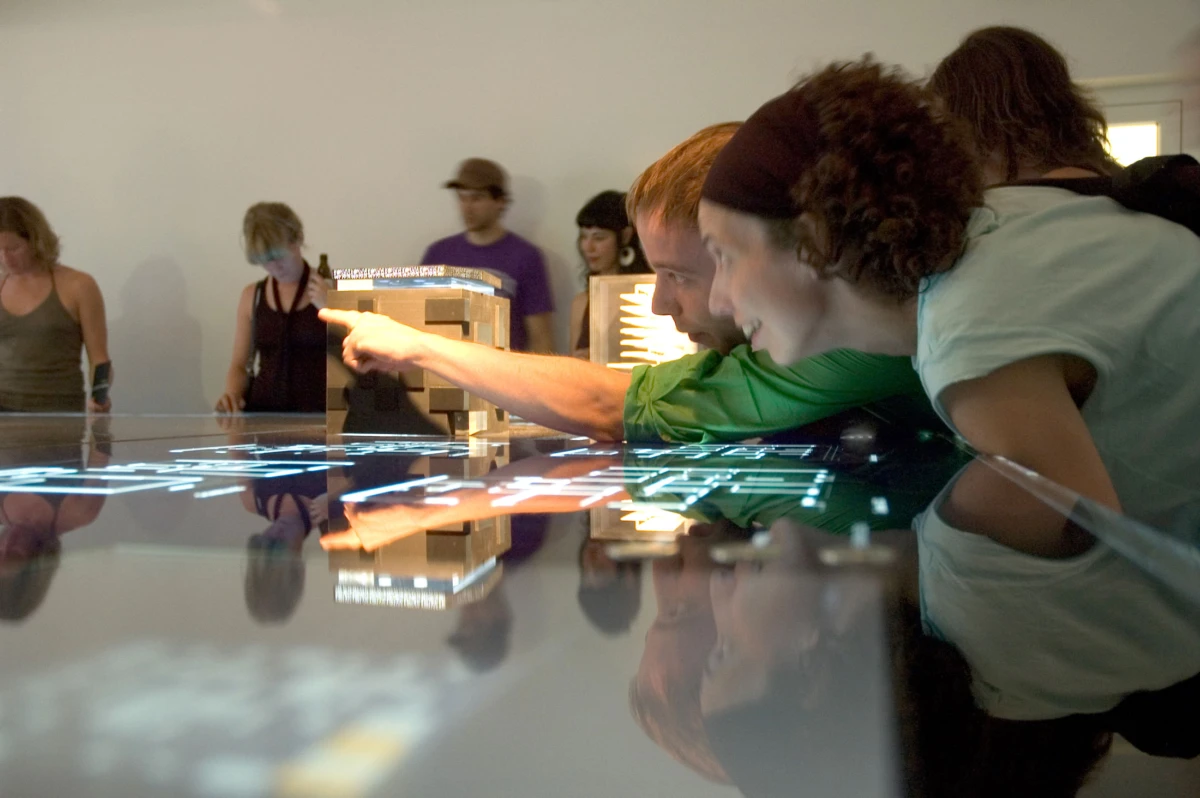Two visitors lean over an interactive touchscreen table, one pointing at the glowing touchscreen interface; onlookers stand in a gallery-like room.