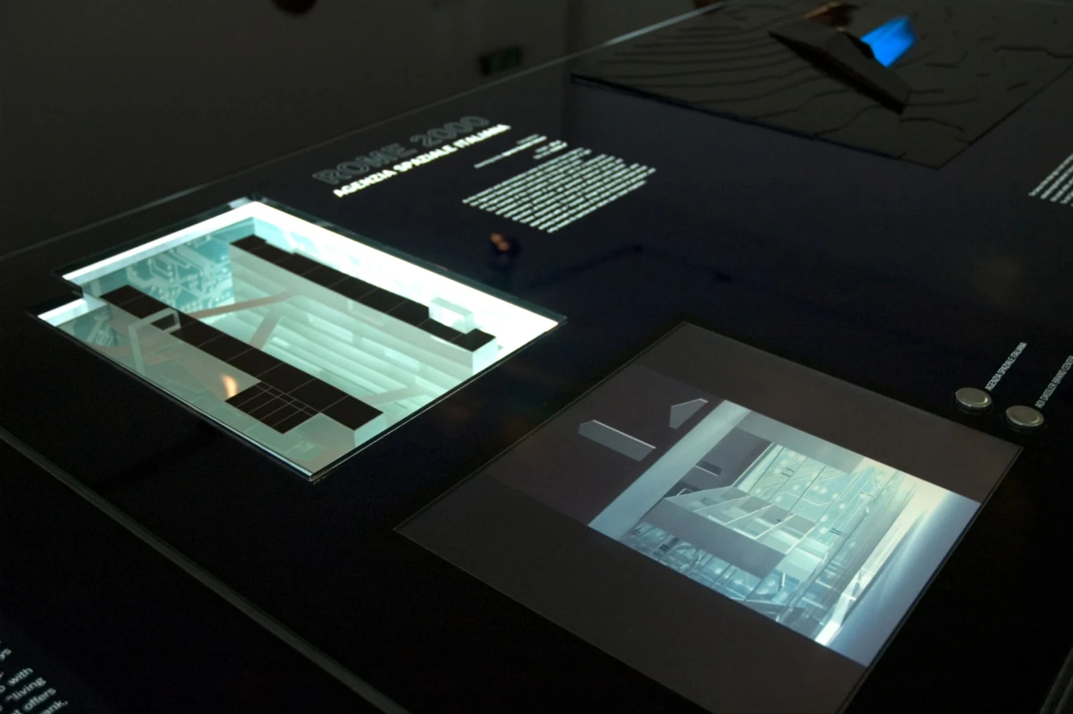 Dark museum display table with a lighted glass-encased architectural model showing a multi-level stairwell; a framed interior photo sits beside it.
