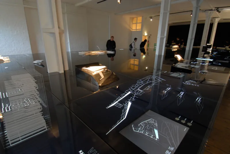 Contemporary art installation on a mirrored black tabletop; geometric wireframe sculptures as visitors observe in a white-walled gallery.