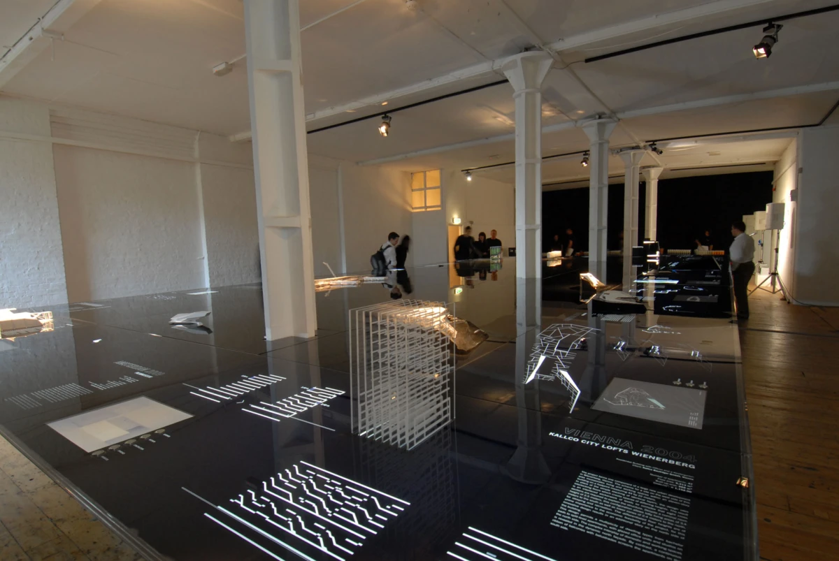 Modern gallery with white columns; reflective black tables display architectural models and text panels, while visitors inspect the exhibits.