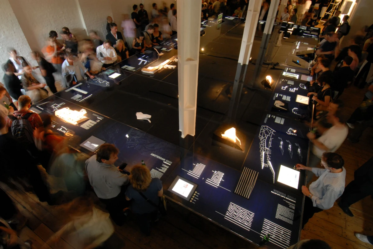 Visitors viewing architectural design exhibits on long display tables in a busy gallery
