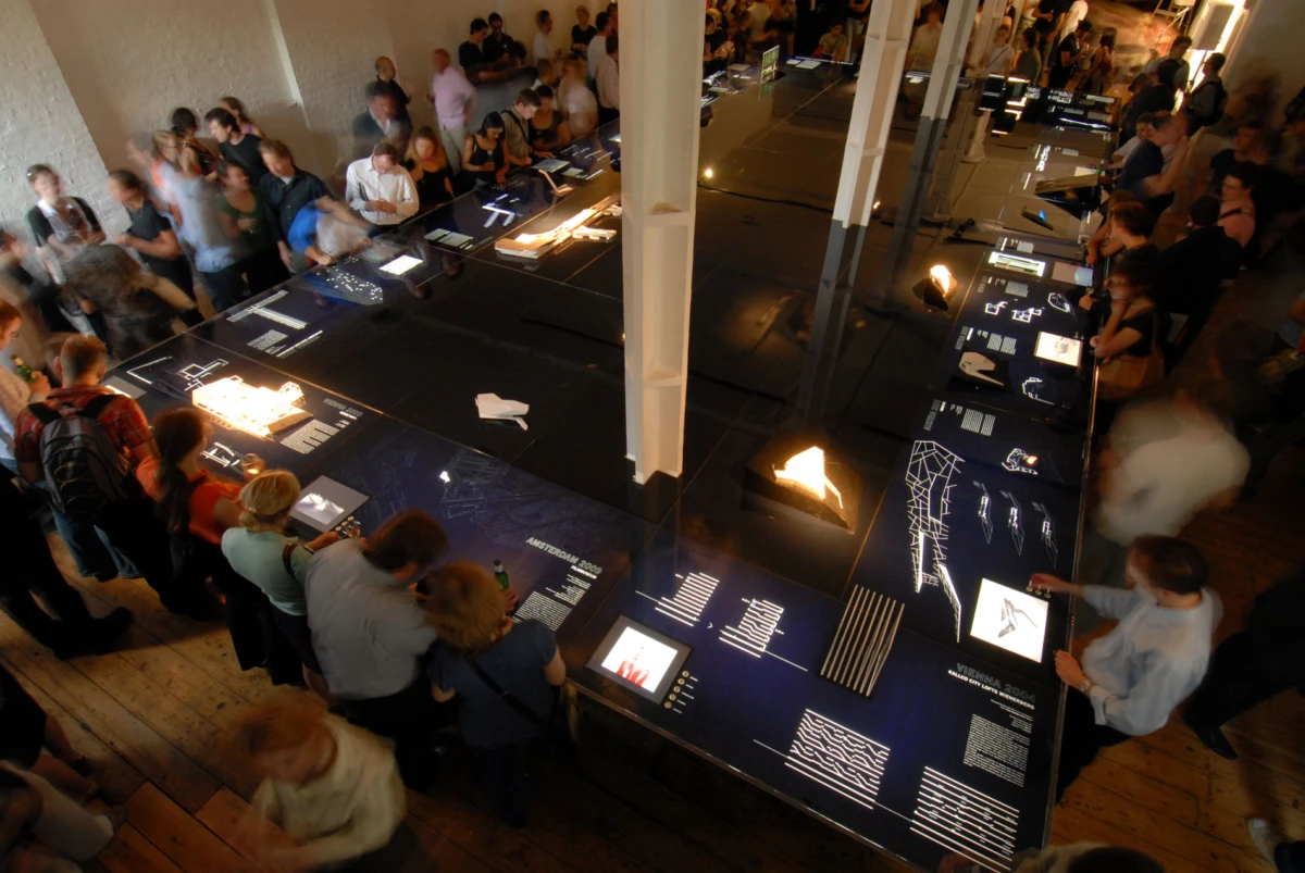 Crowded exhibition hall with long blue display tables and illuminated panels; visitors examine exhibits.