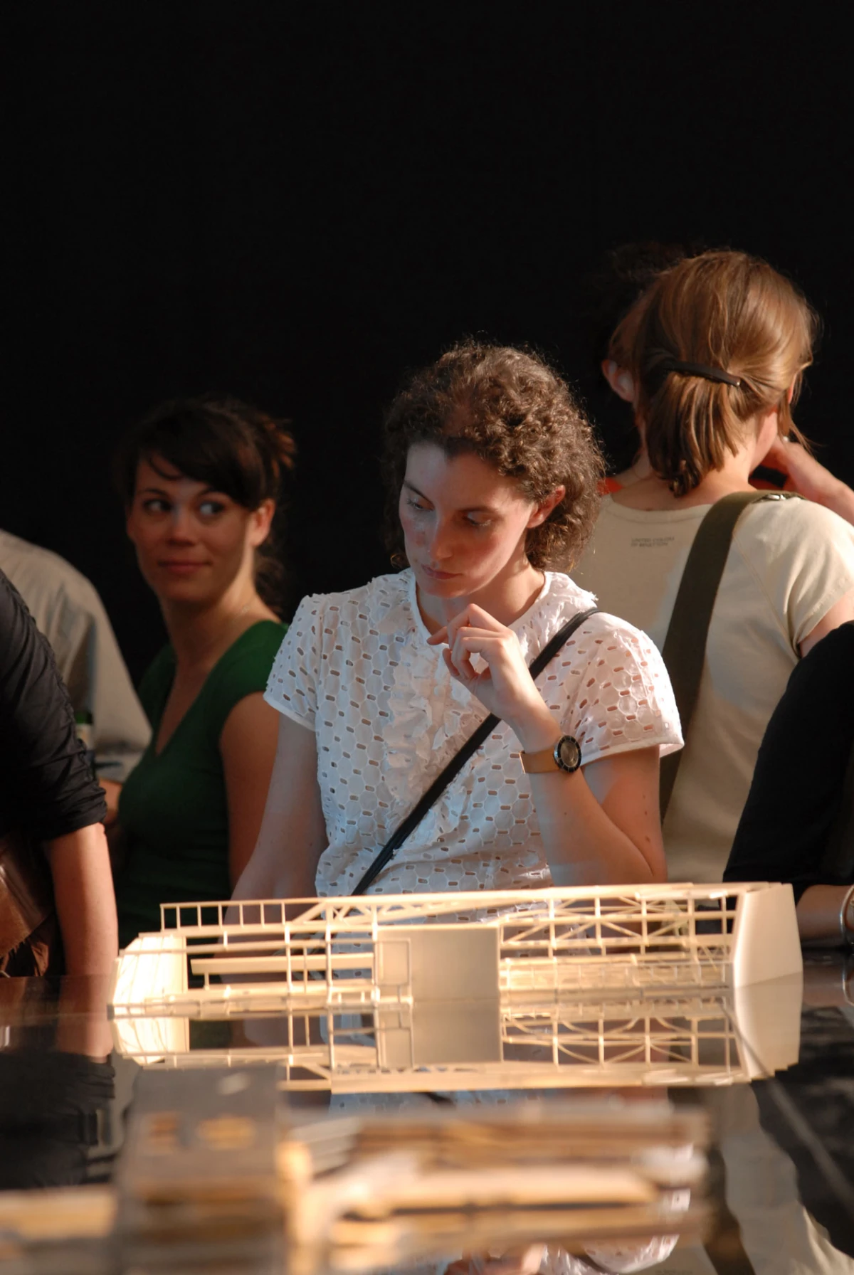 Curly-haired person in a white lace blouse inspects a white architectural model on a glass display table at an exhibition.