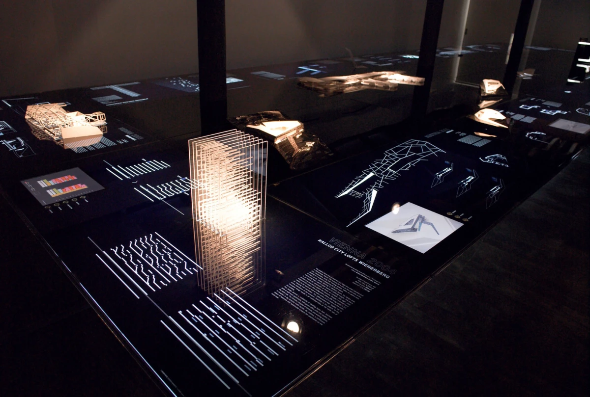Dim museum exhibit with a tall illuminated glass tower model on a glass display, surrounded by white-line architectural diagrams and smaller models.