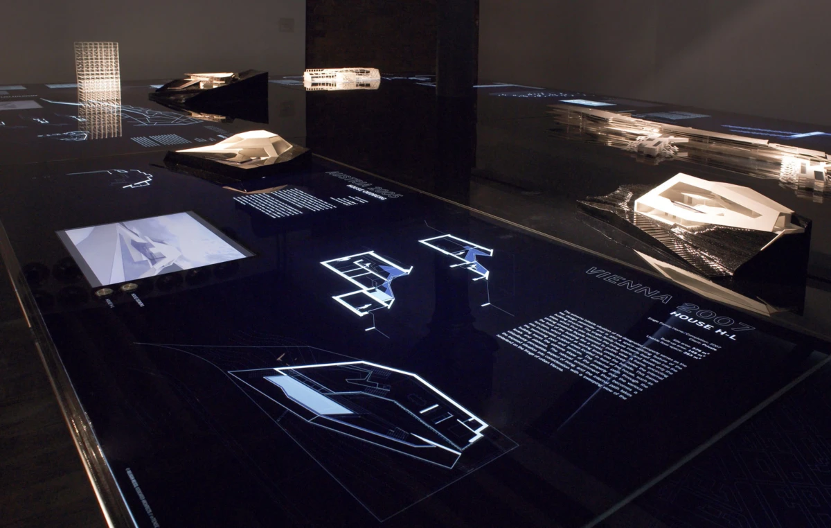 White architectural models displayed on a reflective dark exhibition table, with blueprints and illuminated diagrams in a dim gallery.