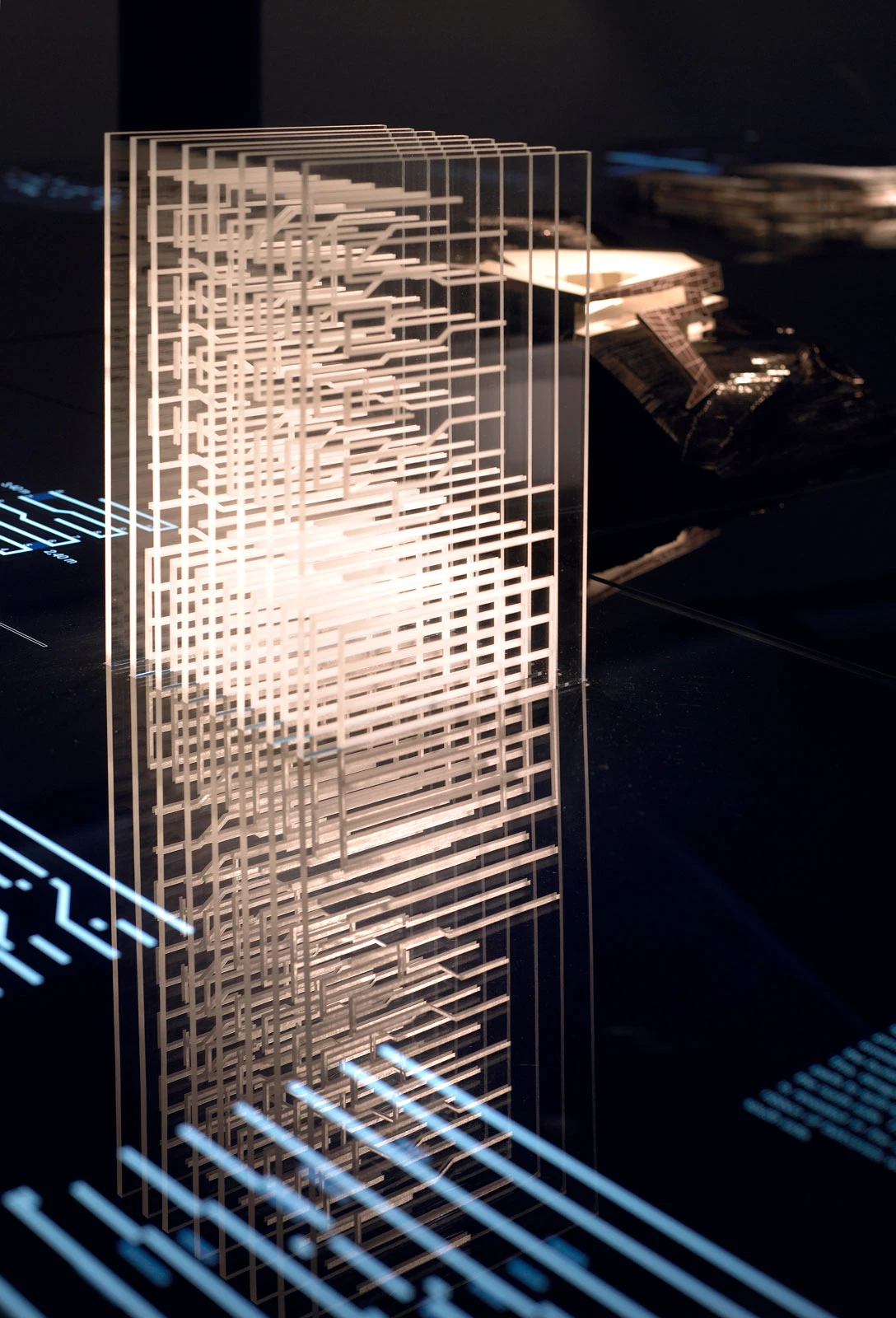 Transparent rectangular sculpture with a layered grid pattern and inner glow, in a dark exhibition space.