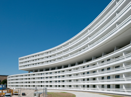 Curved white multi-story apartment building with evenly spaced balconies under a blue sky; construction vehicles at ground level.