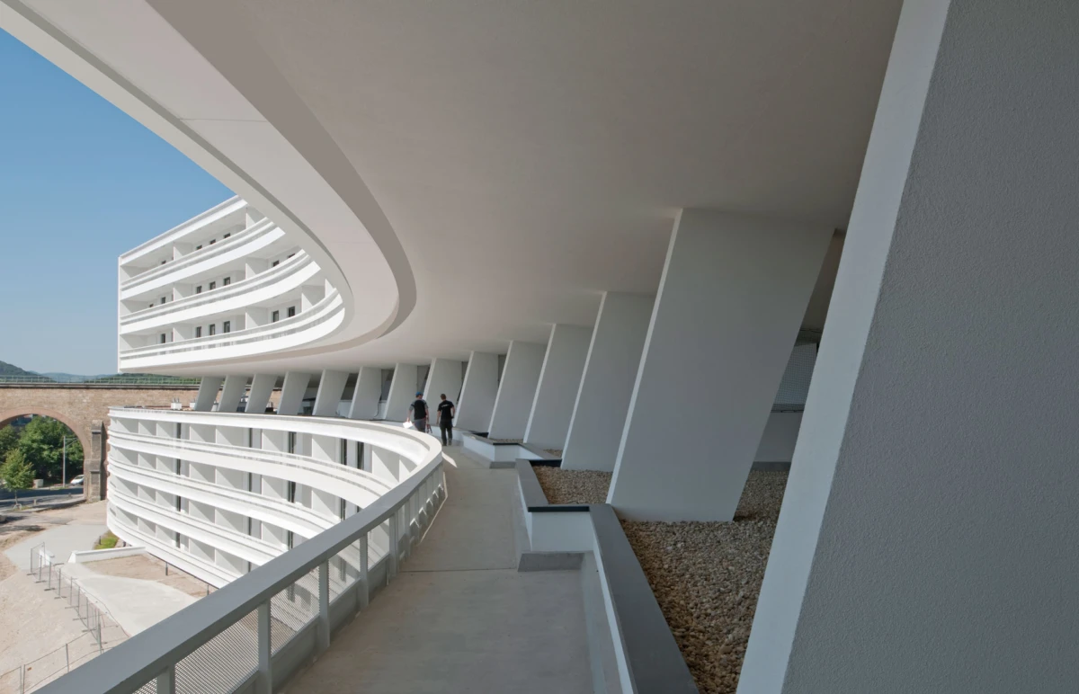 Two people walk along a curved white walkway of a modern building with slanted columns on a sunny day.