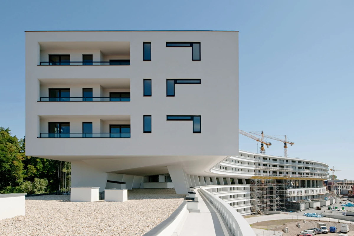 White cantilevered apartment building with three stacked balconies, beside a curved, under-construction structure with cranes in the background.