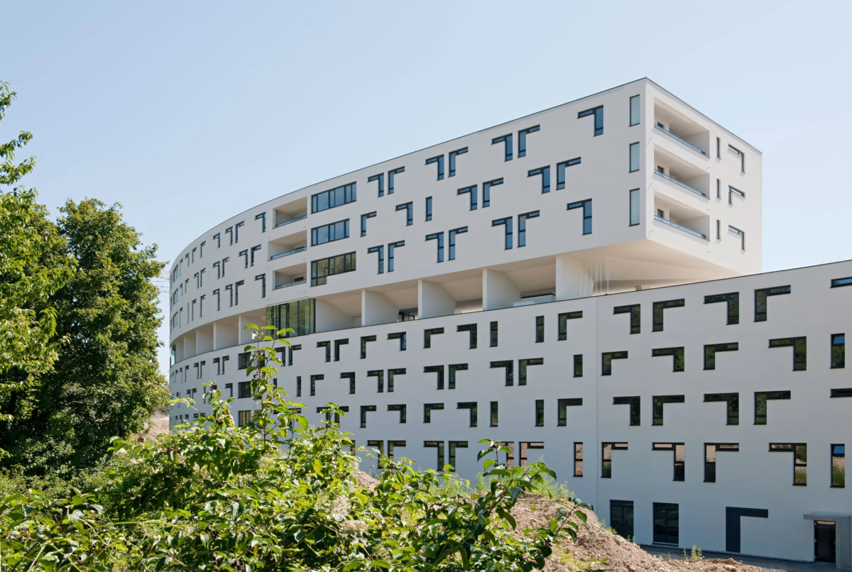 Modern white apartment building with geometric window cutouts and curved facade, set against blue sky with foreground greenery.