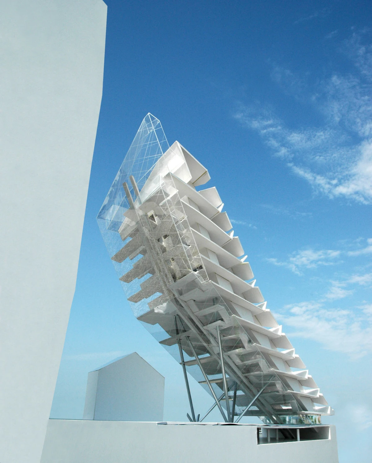 Tilted white architectural sculpture with stacked fins on metal supports atop a rooftop, glass-lattice tip against blue sky