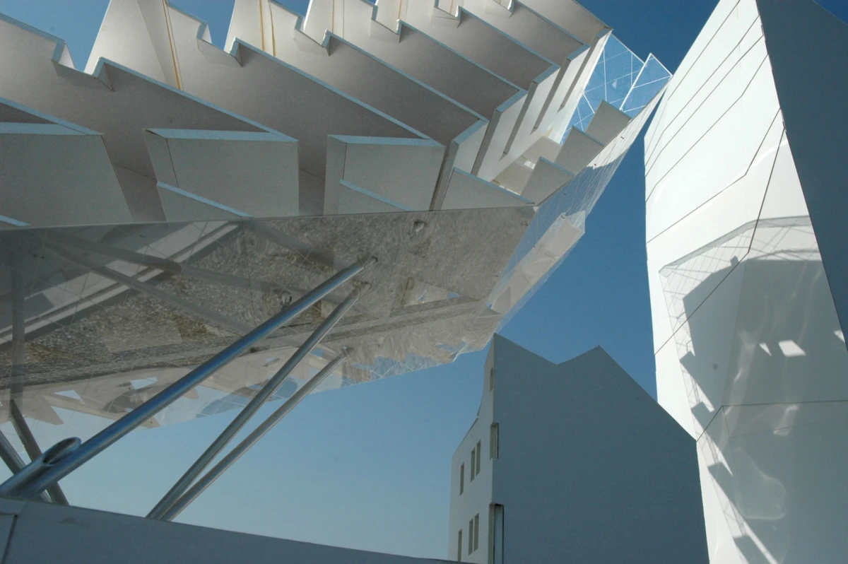 Angular white building facade with glass railing and steel support rods against a blue sky.