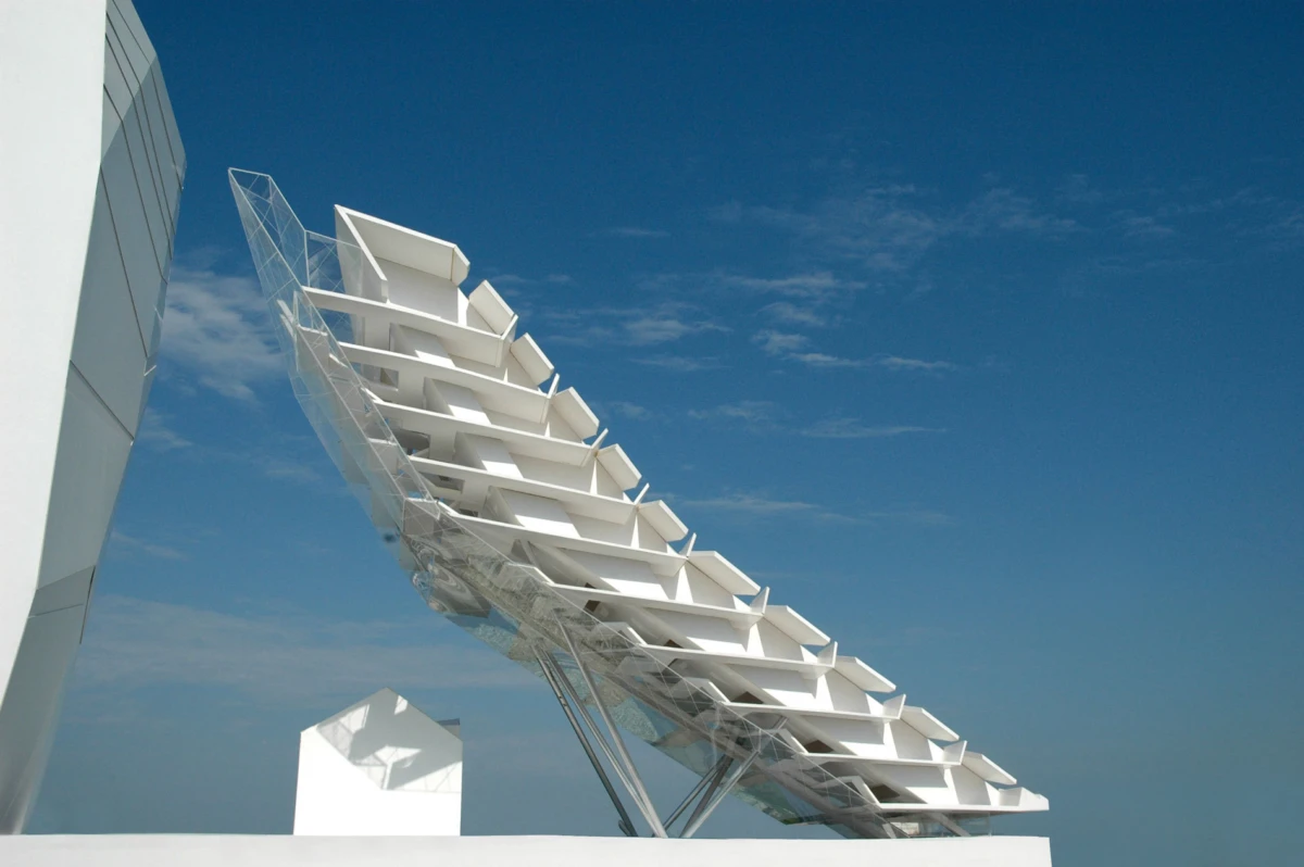 White cantilevered staircase sculpture supported by metal framework against a clear blue sky.