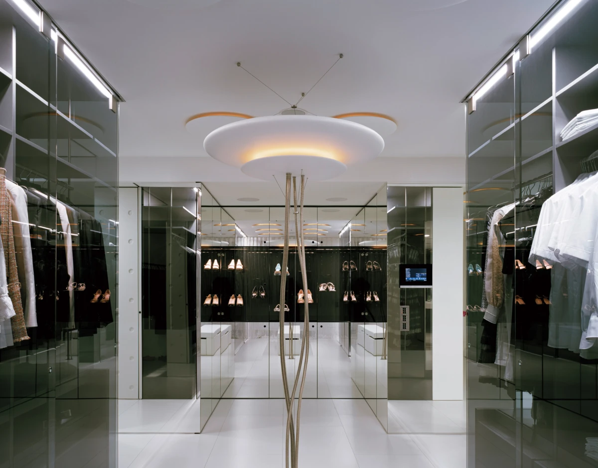 Modern fashion boutique interior with glass display walls, clothing and shoe racks, and a sculptural circular ceiling light.