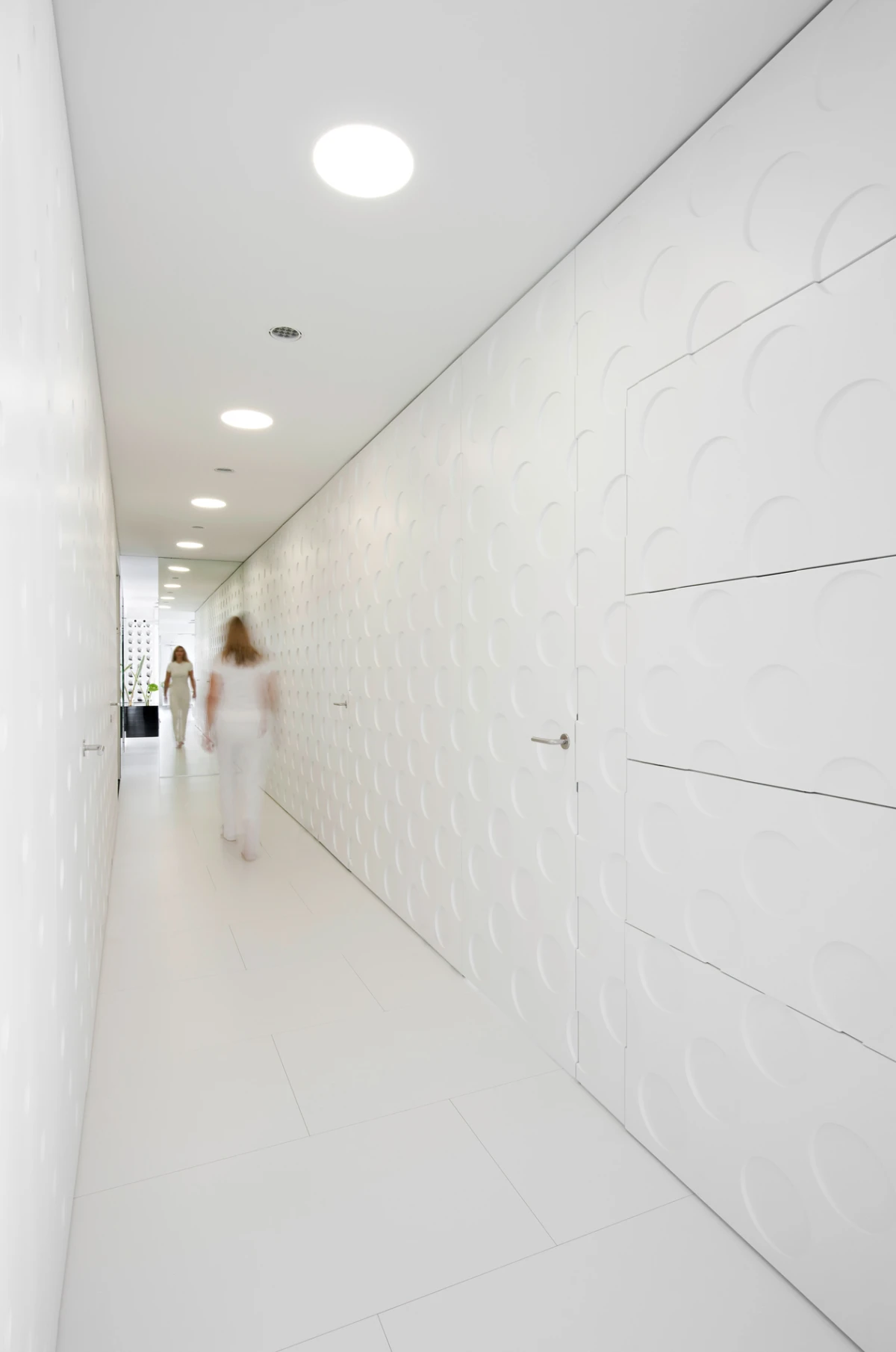 Two individuals in white outfits walking away in a white minimalist corridor lined with circular wall panels.