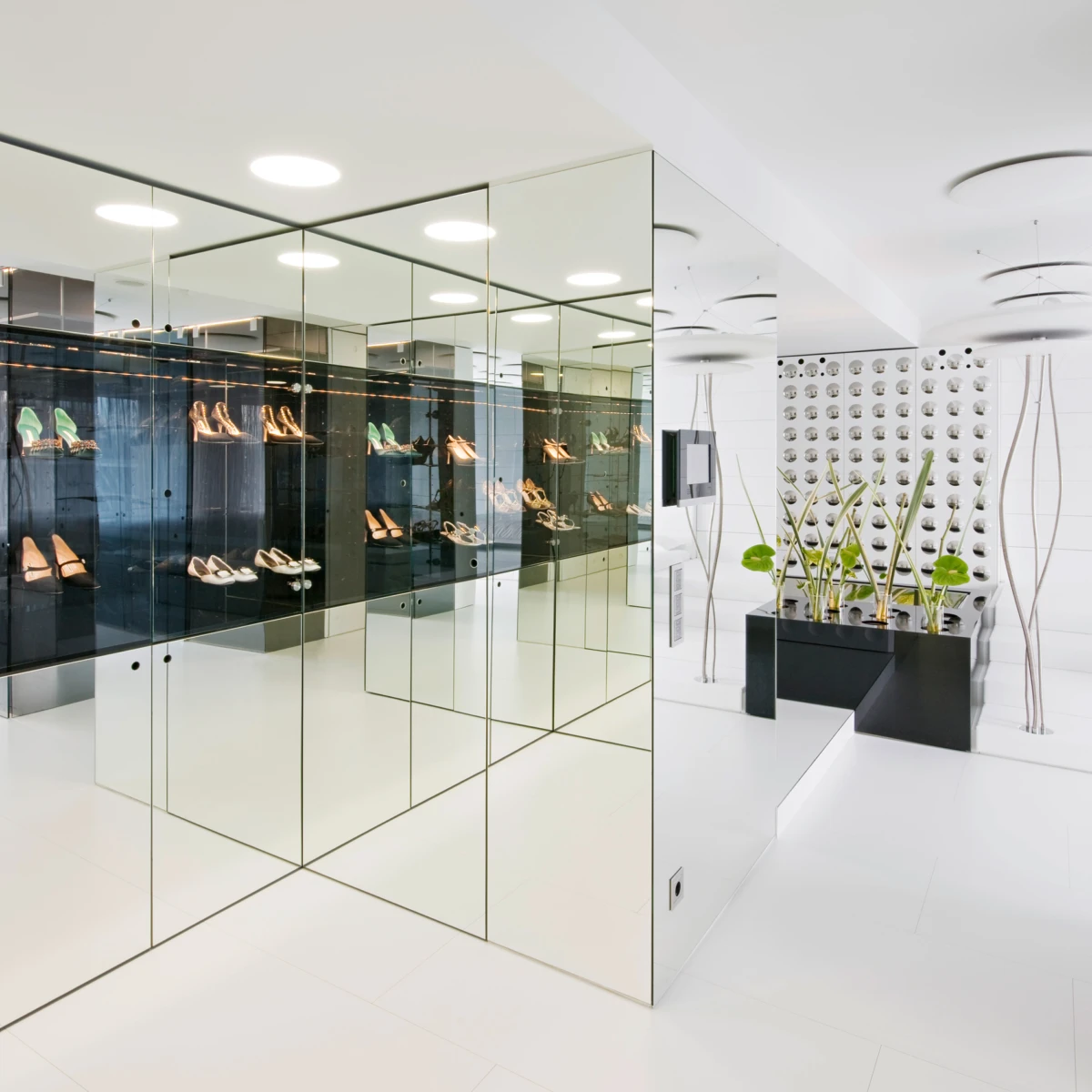 Minimalist shoe showroom with mirrored glass display cabinets showcasing high-heeled shoes; white floor, black planters, and tall decorative plants.